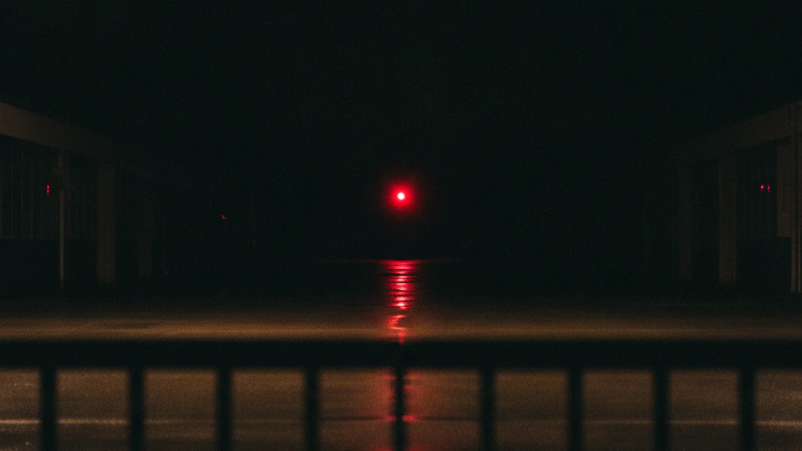Pátio vazio à noite, asfalto molhado refletindo uma luz vermelha ao fundo, cerca metálica desfocada em primeiro plano