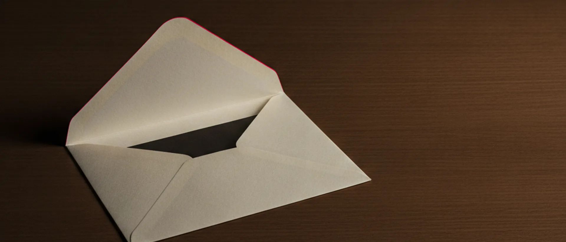 Envelope off-white vazio e aberto sobre mesa de madeira escura, com o interior em sombra e um fio de luz vermelha na aba