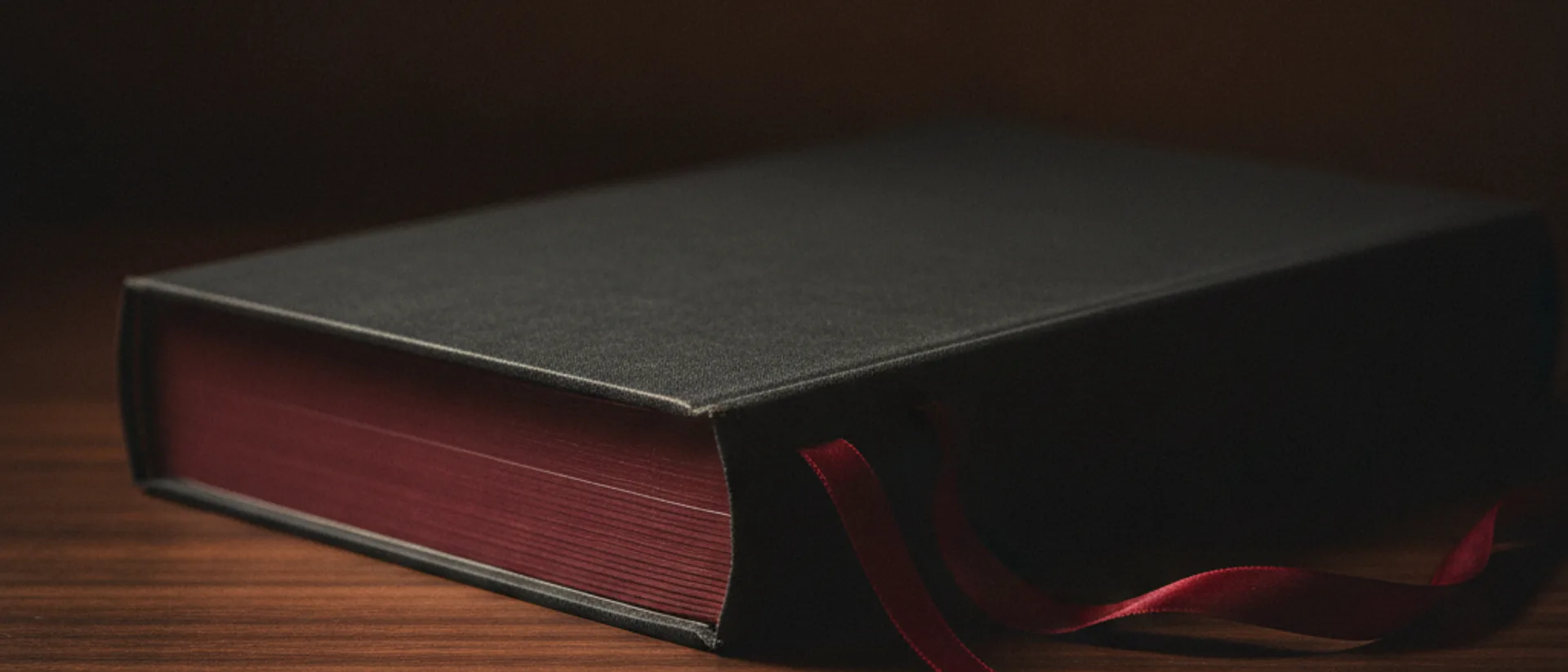 Livro de capa dura fechado sobre mesa escura, com fita marcadora vermelha saindo entre as páginas