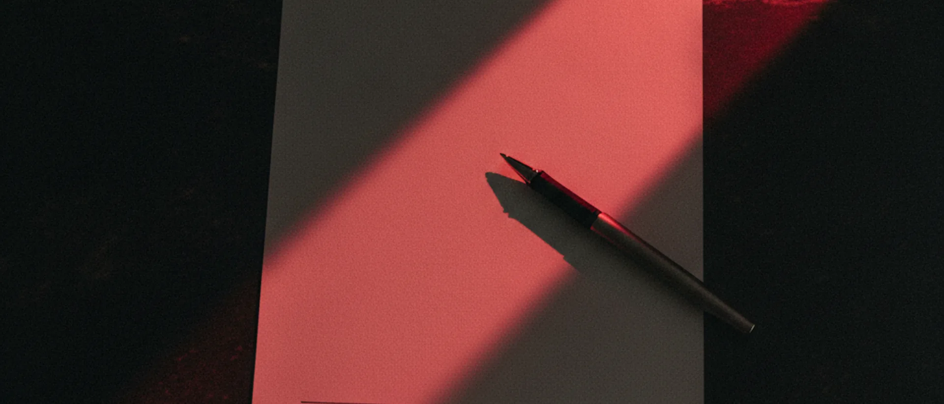 Contrato de papel sobre mesa escura com uma caneta pousada perto da linha de assinatura, cortado por uma faixa de luz vermelha